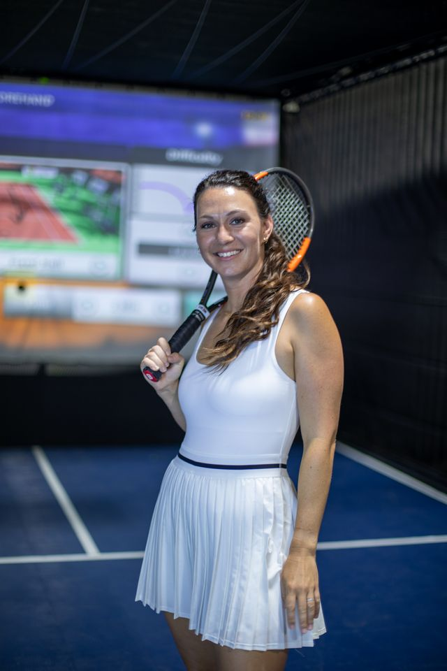 A woman holding a tennis racket and smiling at the camera, standing on a tennis simulator court with tennis courts and a tennis simulator in the background.