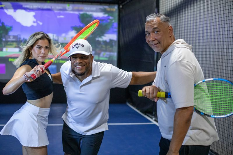 Three people at an indoor tennis simulation facility, holding tennis rackets, smiling, with a large screen displaying a virtual tennis court in the background.