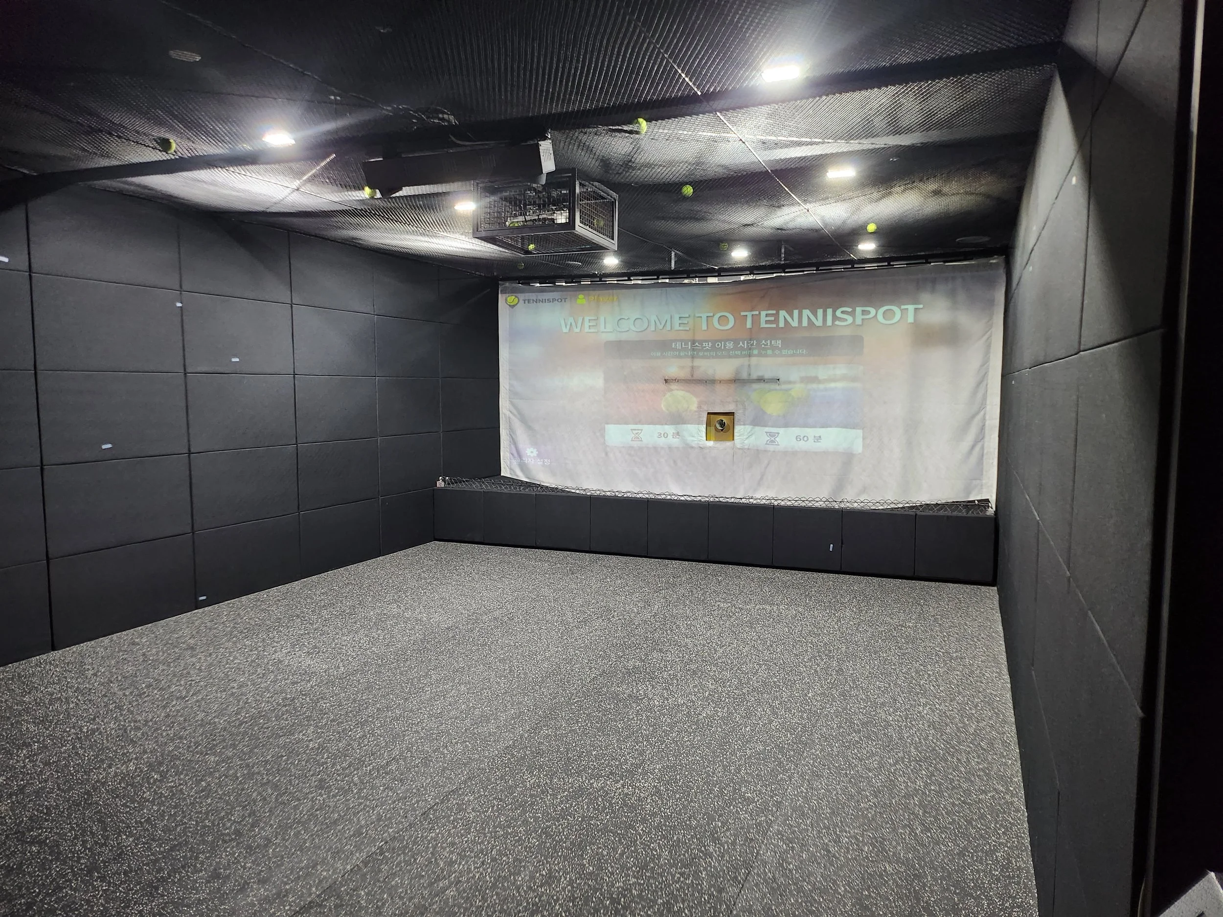 Indoor tennis practice area with a large screen displaying the message 'Welcome to Tennispot' and a tennis ball vending machine near the wall.