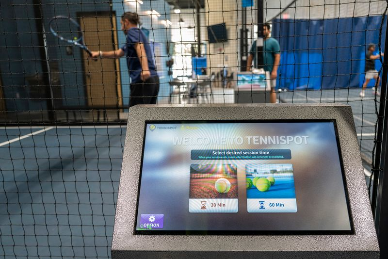 Tennis court indoors with players practicing; touchscreen kiosk in the foreground showing session time options of 30 or 60 minutes.