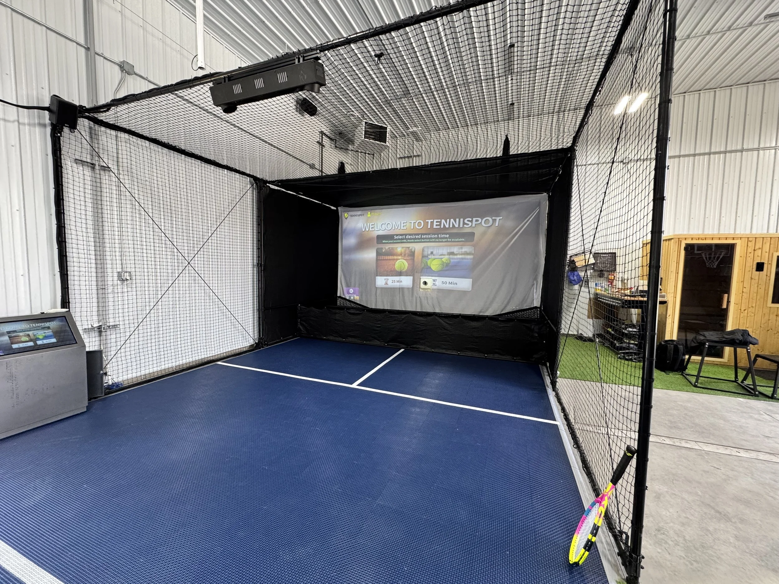 Indoor tennis hitting cage with blue court surface, black netting, and a welcome sign for Tennispot with session options of 25 or 55 minutes.