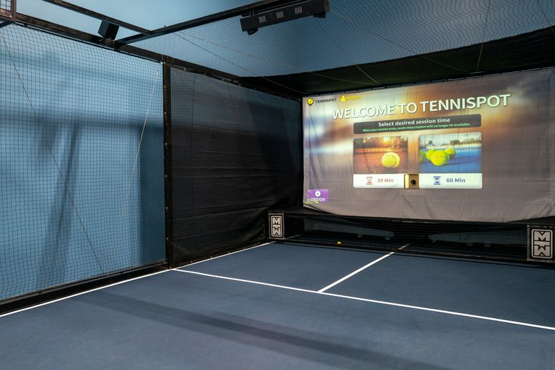 Indoor tennis simulator with a large screen displaying a tennis-themed welcome message and session options for 30 or 60 minutes.