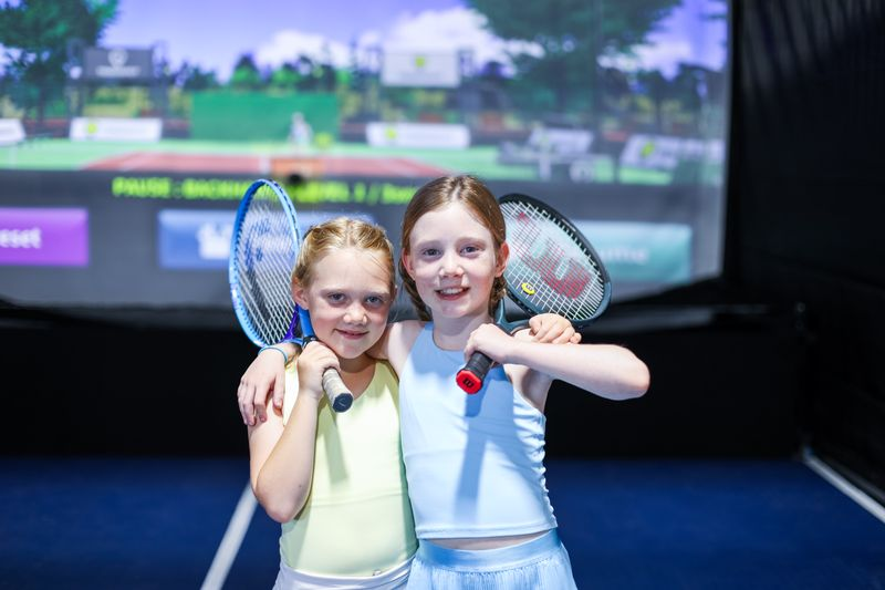 Two young girls with tennis rackets smiling at a tennis simulation game