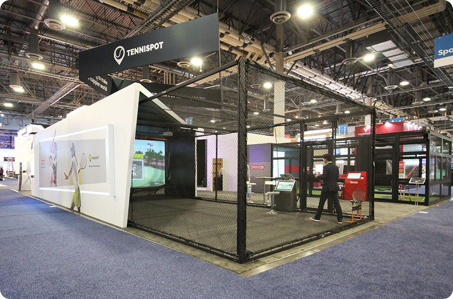An indoor exhibition booth for Tennis Spot at a convention center, featuring a large tennis-related digital screen and a batting cage for tennis practice, with people inside and a visible overhead sign displaying the Tennis Spot logo.