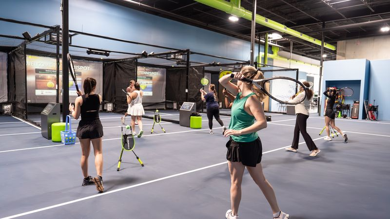 People practicing tennis indoors at a tennis facility with multiple hitting stations and screens.