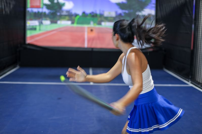 Young woman playing tennis indoors on a tennis simulator, hitting a tennis ball with her racket, with a large screen in the background displaying a tennis court.