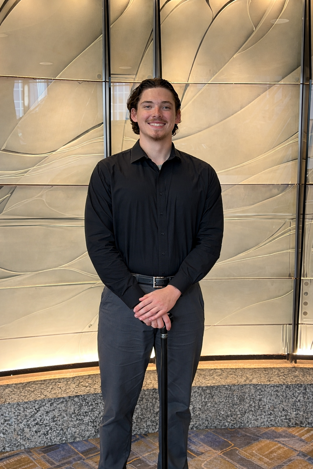 A young man with dark, wavy hair, wearing a black button-up shirt and gray pants, standing in front of an abstract decorative wall with gold accents, smiling at the camera.