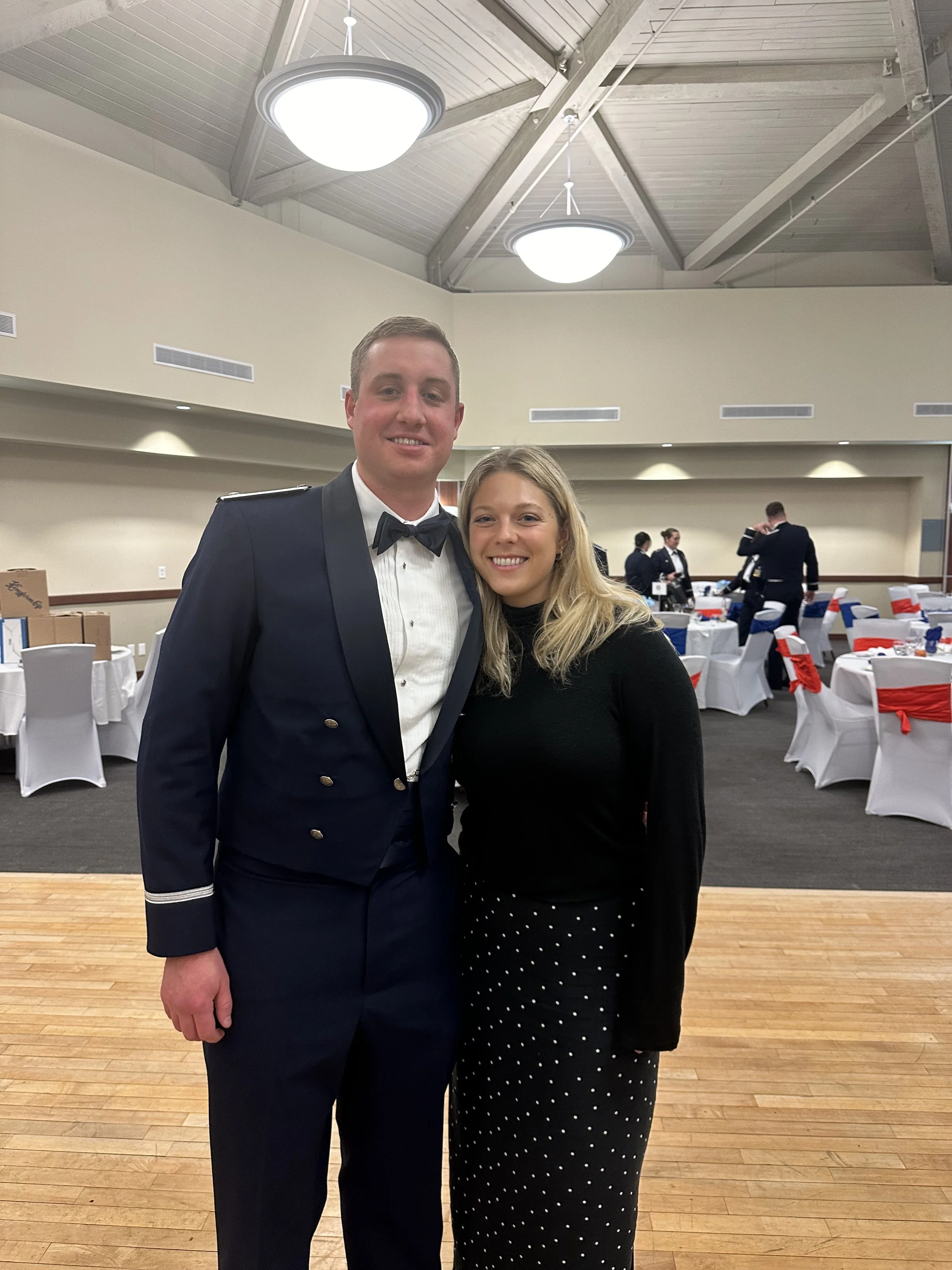 A young man in a navy blue military uniform and a young woman in black and white polka dot pants and a black top standing together in a banquet hall with decorated chairs and people in the background.