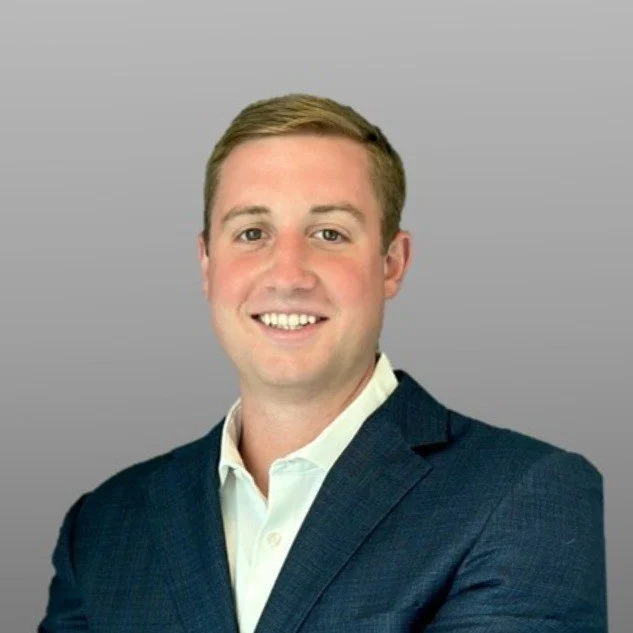 A young man with short brown hair and fair skin, wearing a dark blue suit jacket and a white shirt, smiling at the camera against a plain gray background.
