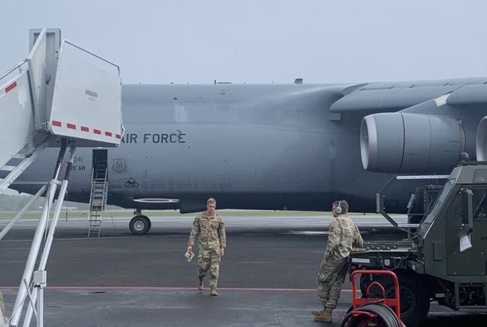 Two military personnel in camouflage uniforms walking near a large cargo plane on an airport tarmac, with equipment and stairs nearby.