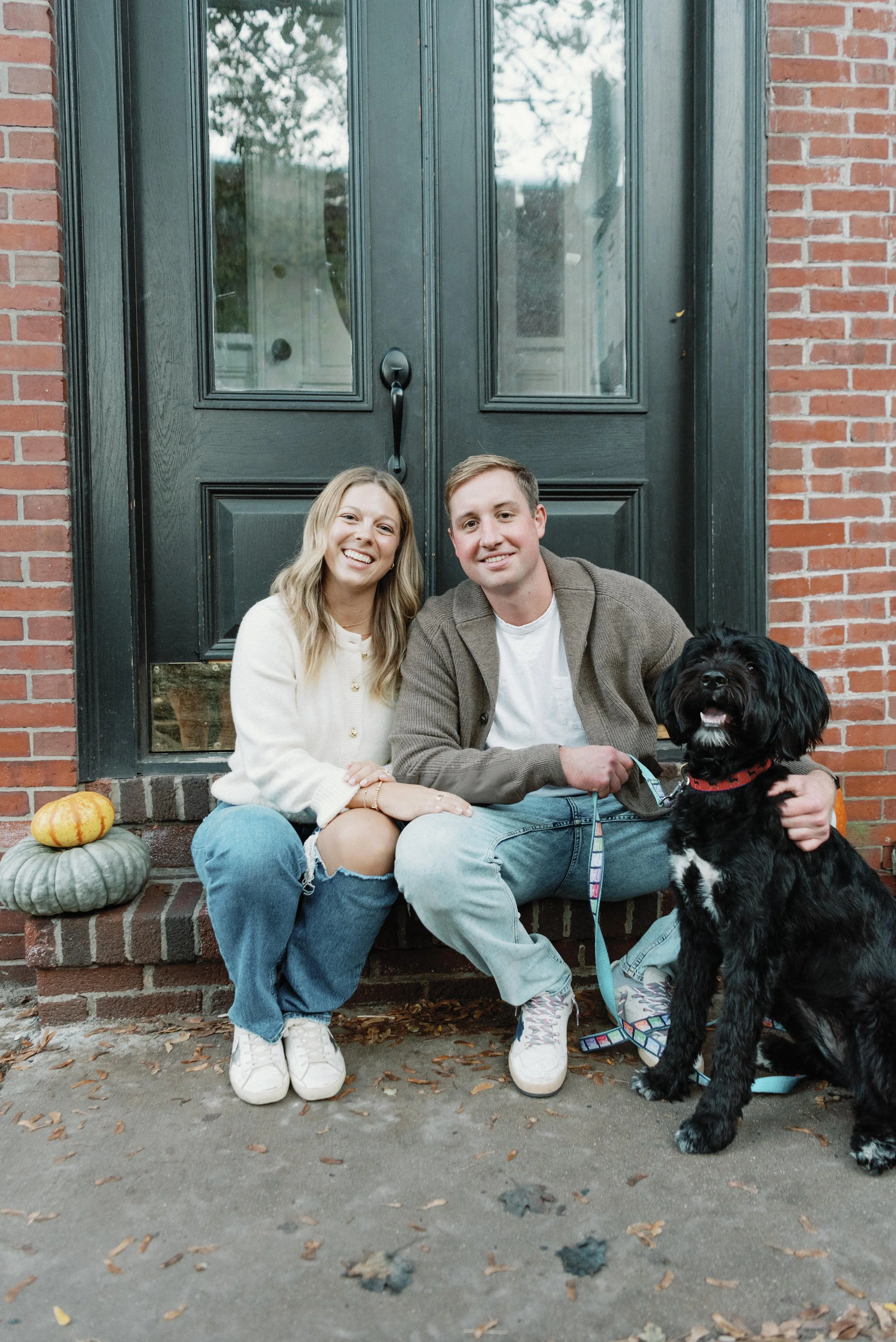 A smiling young couple sitting on steps in front of a black door, with a black dog, pumpkins, and yellow leaves around them.