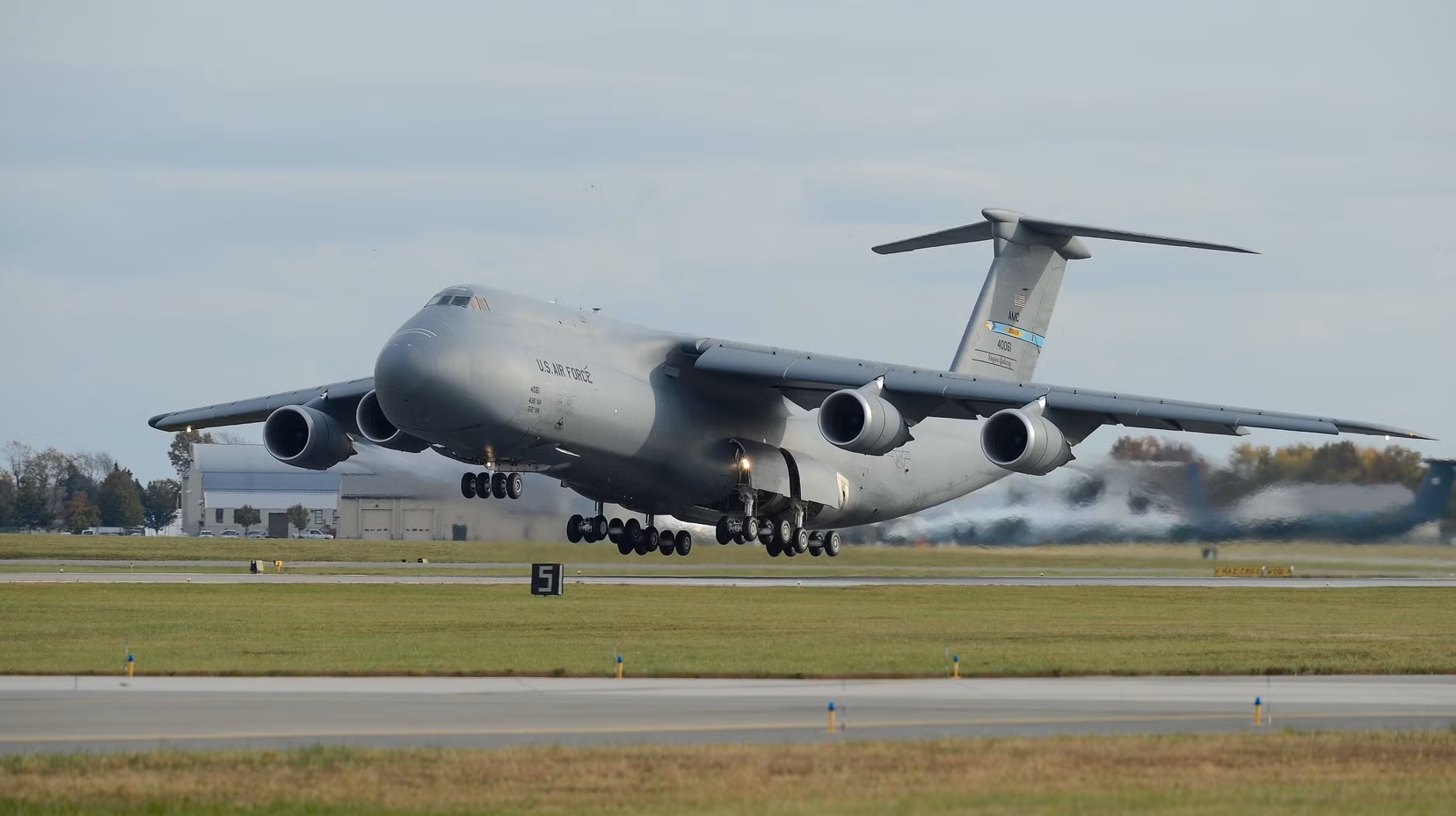 A large gray military cargo jet plane taking off from an airport runway with smoke trailing behind engine exhaust.