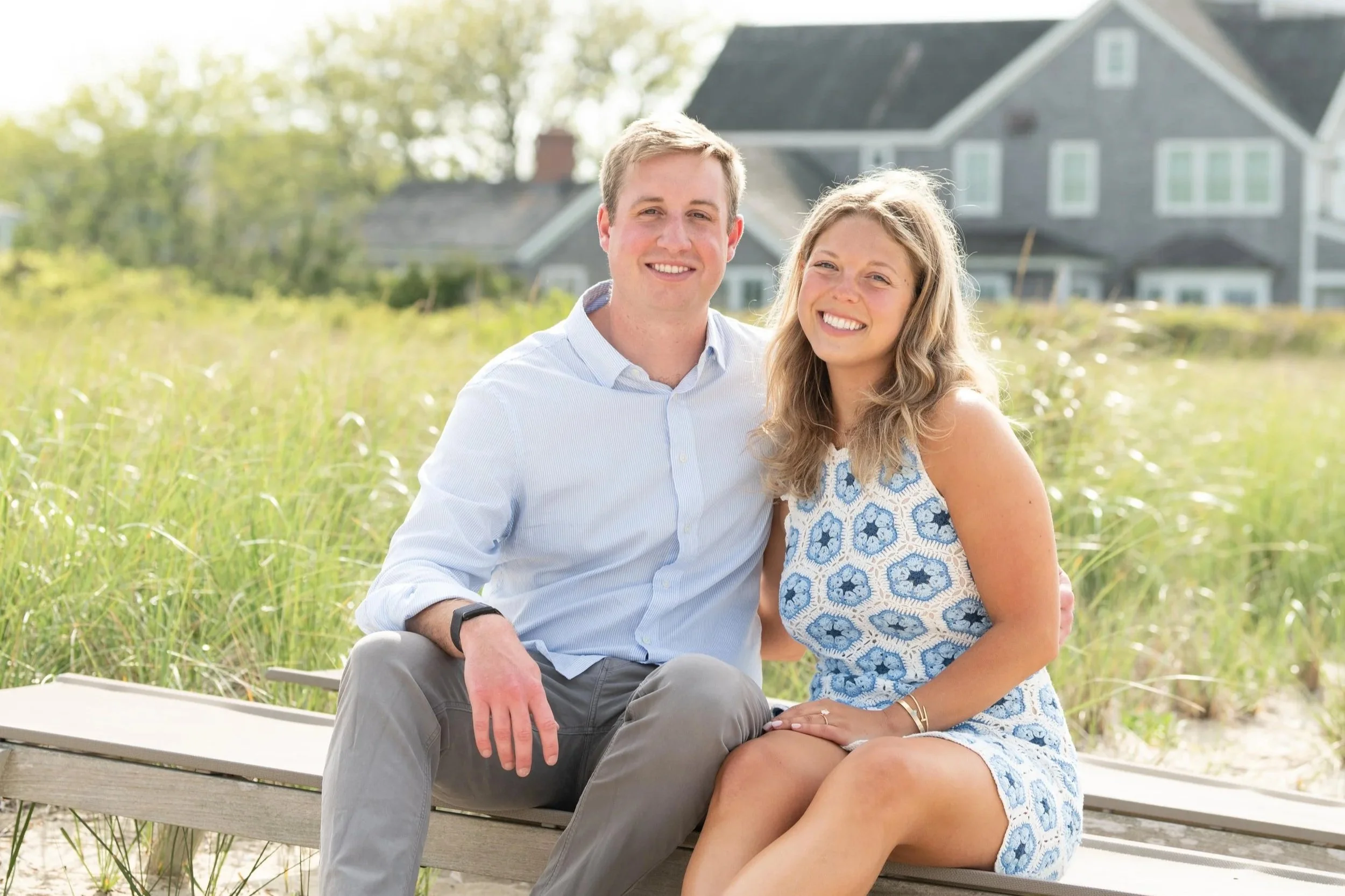 A young man and woman sitting closely on a bench outdoors, smiling at the camera, with grassy dunes and houses in the background.