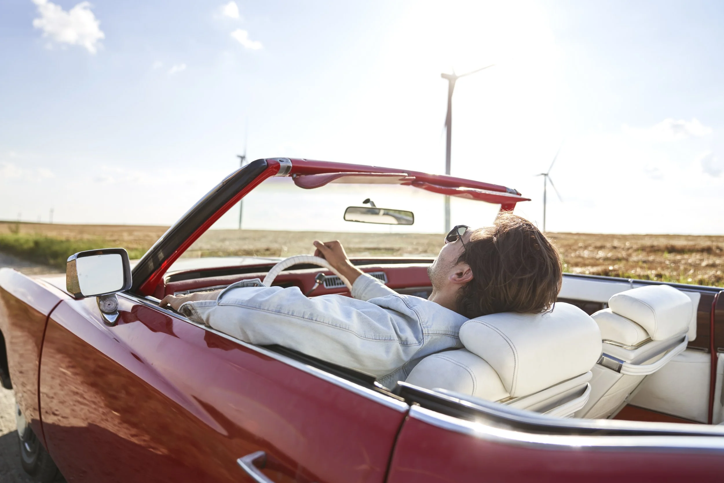 A man lying back in a red convertible car with white interior, resting on the driver's seat with a wind farm in the background during daytime.