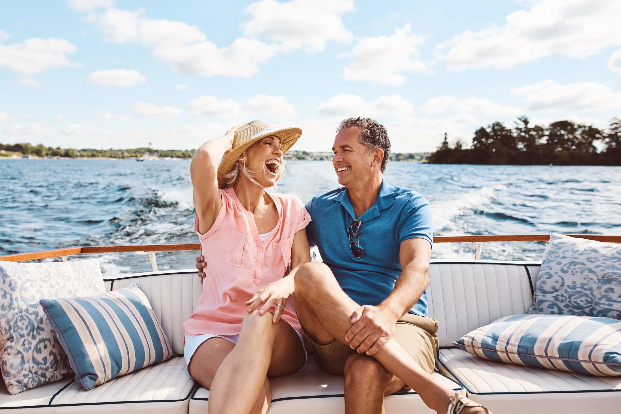 A couple enjoying a boat ride on a sunny day, sitting on a cushioned bench at the back of the boat, surrounded by water and a scenic distant shoreline with trees.