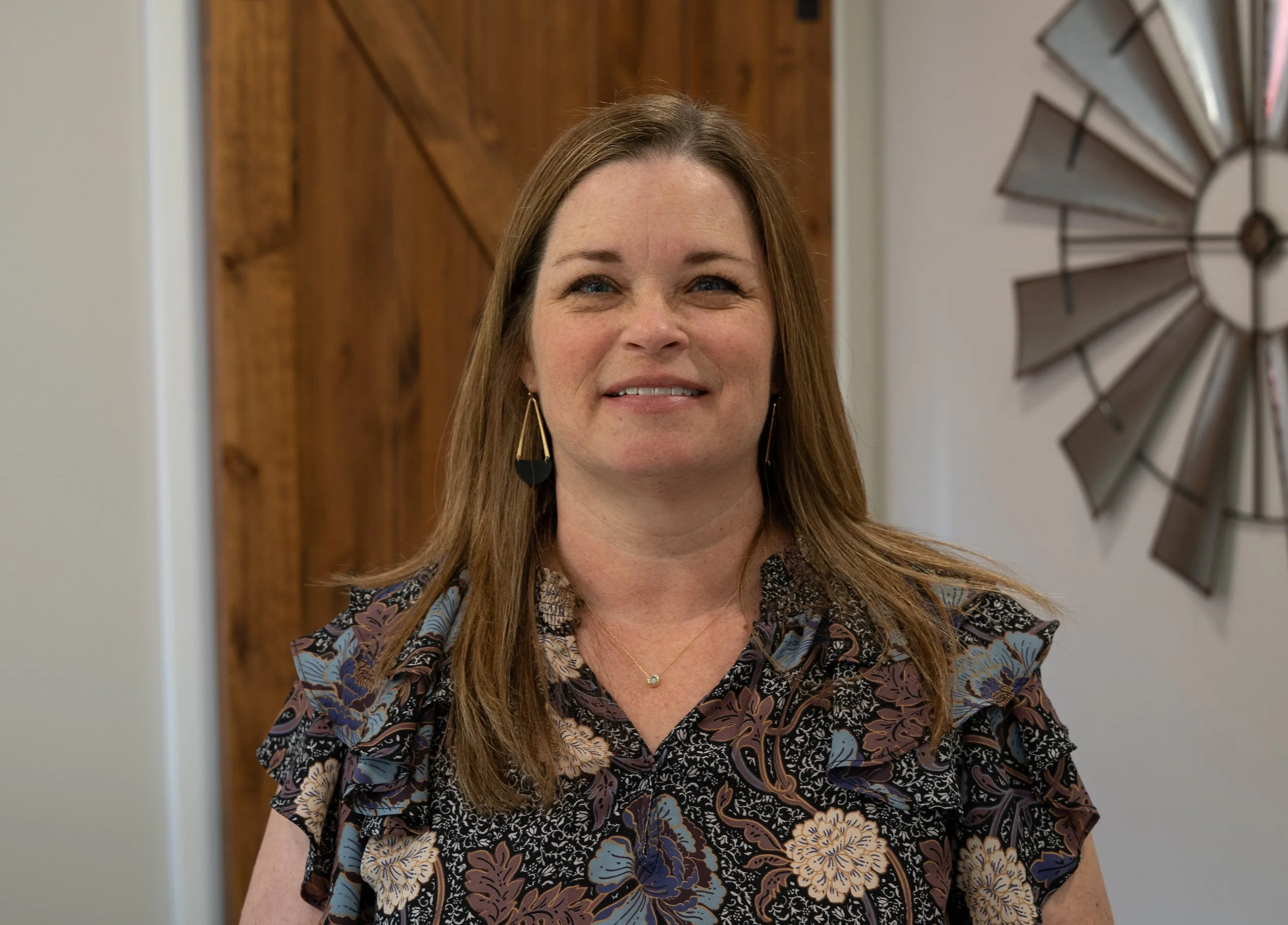 A woman with shoulder-length brown hair, wearing a floral patterned blouse, gold earrings, and a small pendant necklace, standing indoors near a wooden door and decorative wall art.