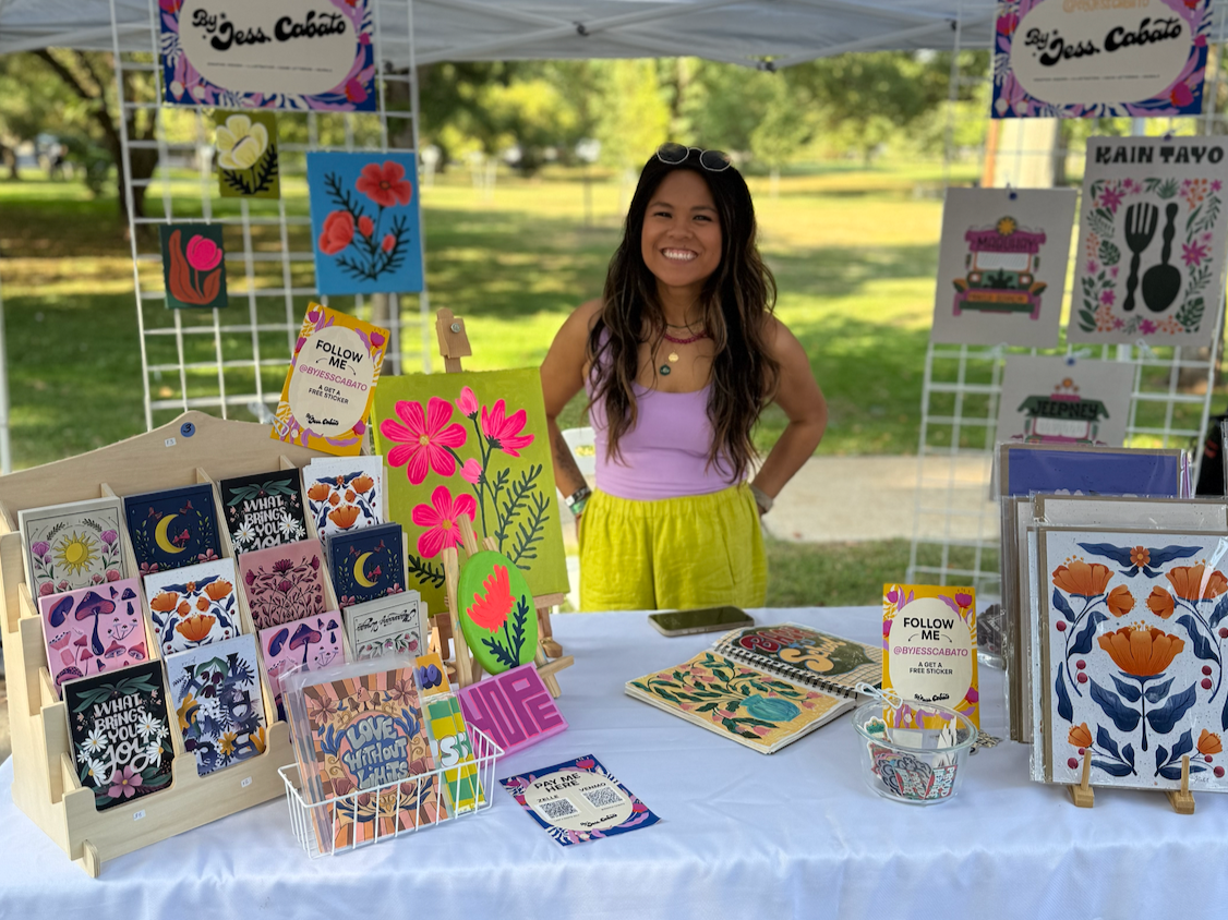Woman standing behind a booth at an outdoor market, displaying colorful floral and abstract art prints and cards.
