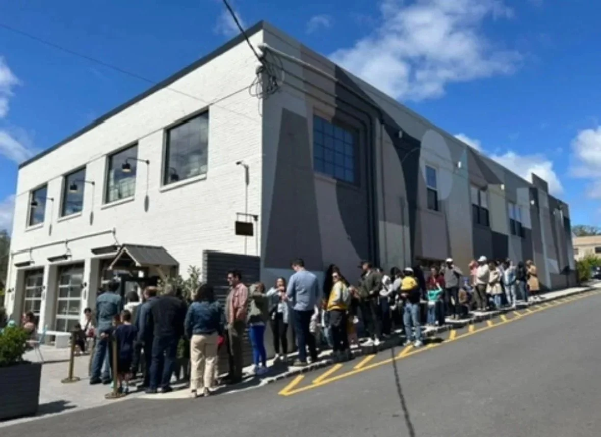 Long line of people waiting outside a modern, multi-story building on a sunny day.