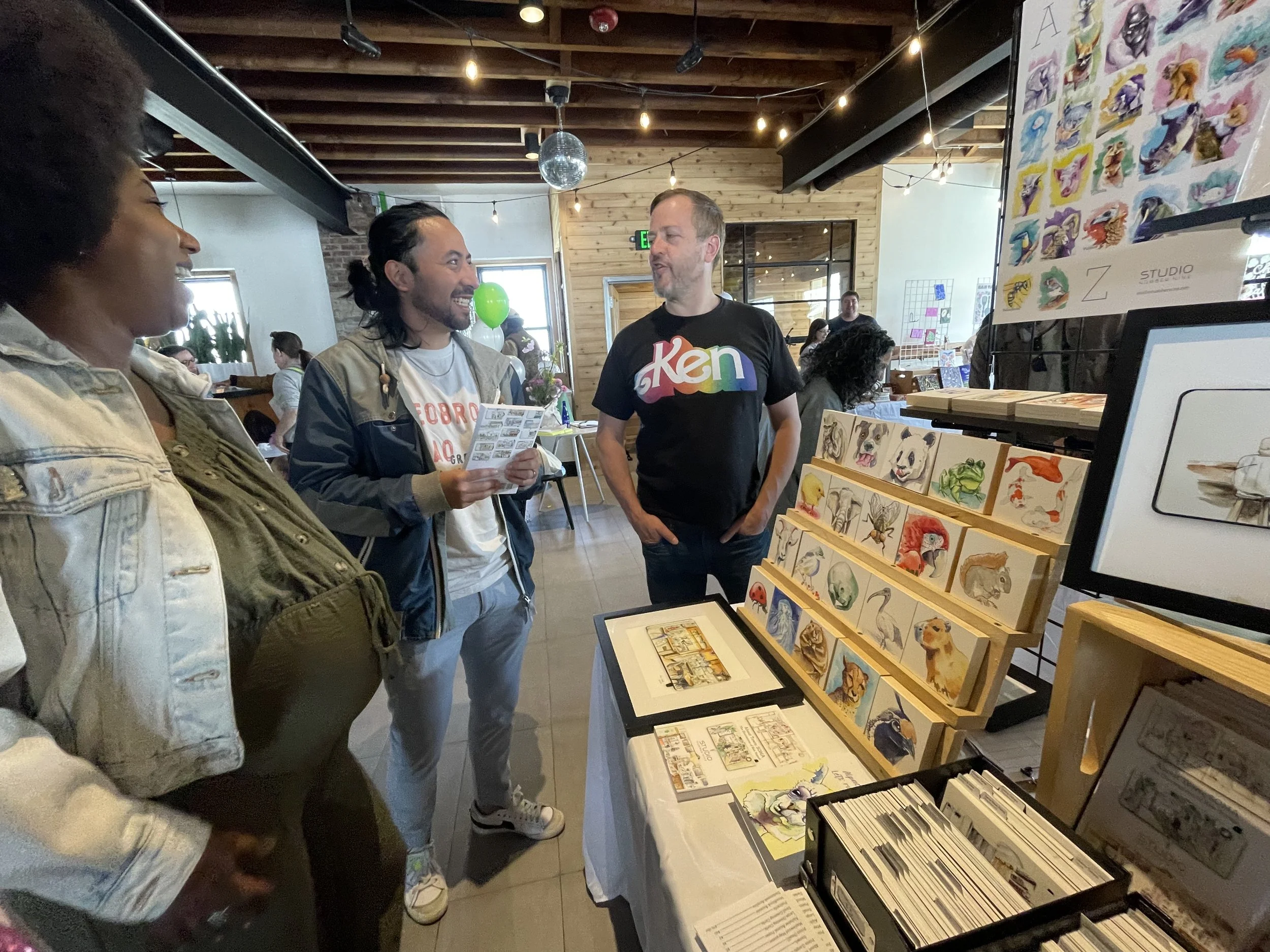Three people are talking at an art gallery or craft fair with various artwork displayed on tables and racks. The background includes other visitors, wooden walls, and string lights.