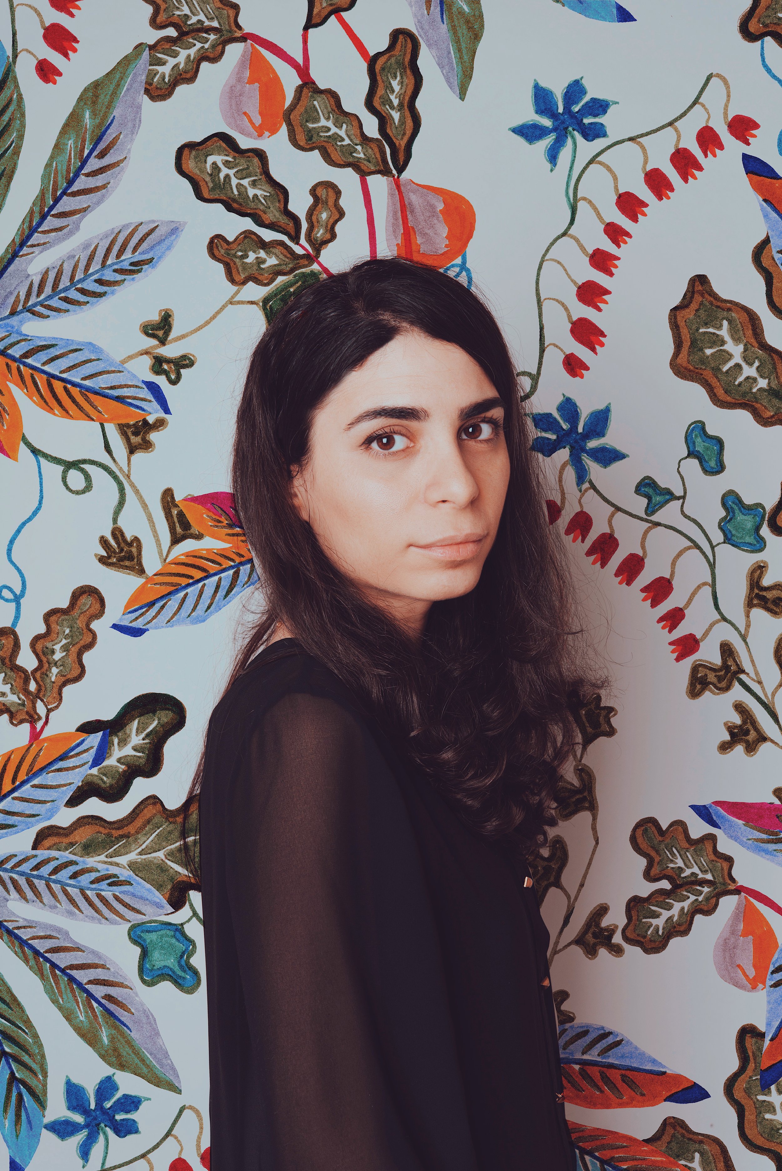 A woman with dark wavy hair and fair skin standing in front of a colorful, botanical-patterned wall.