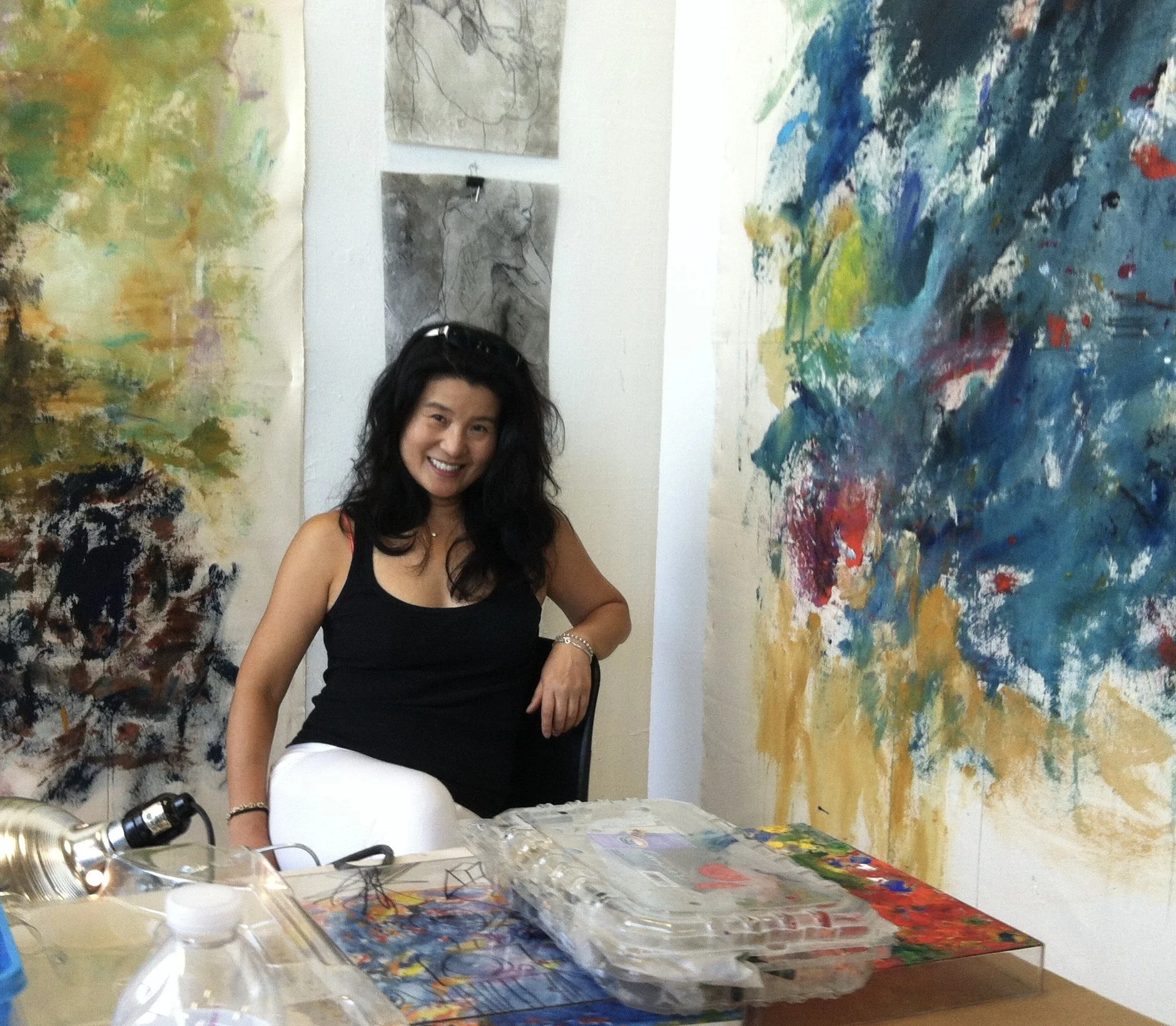 A woman with black hair, wearing a black tank top and white pants, sitting at a table in an art studio surrounded by colorful abstract paintings and sketches.