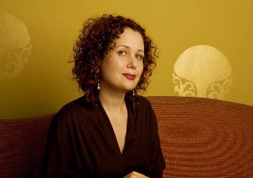Portrait of a woman with curly hair, wearing a black top, sitting in front of a yellow background with abstract patterns.