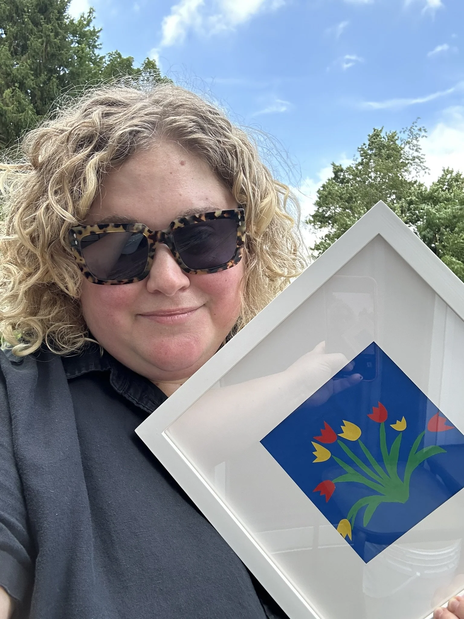 A woman with curly blonde hair, wearing sunglasses and a black shirt, holding a frame with a colorful floral artwork outdoors under a blue sky with clouds and green trees in the background.