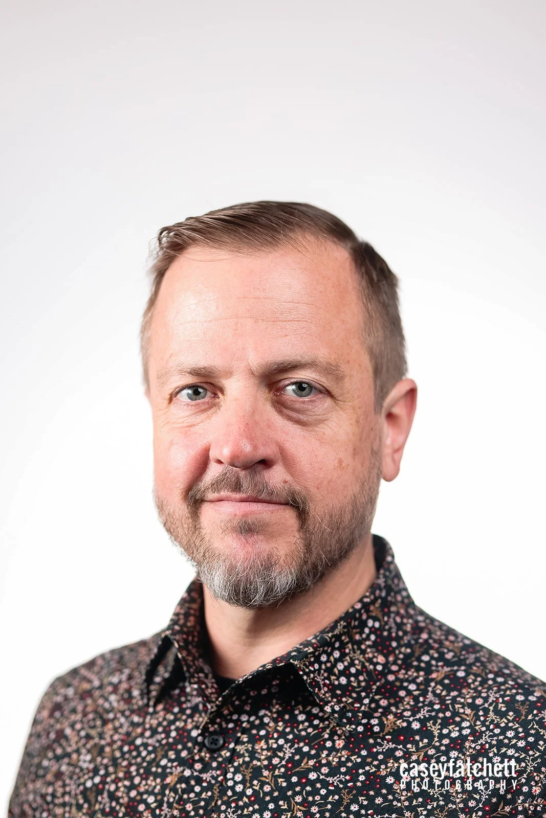 Portrait of a man with short brown hair, a beard, wearing a floral patterned shirt, against a plain white background, with the photographer's watermark 'caseyfatteh.photography' in the bottom right corner.