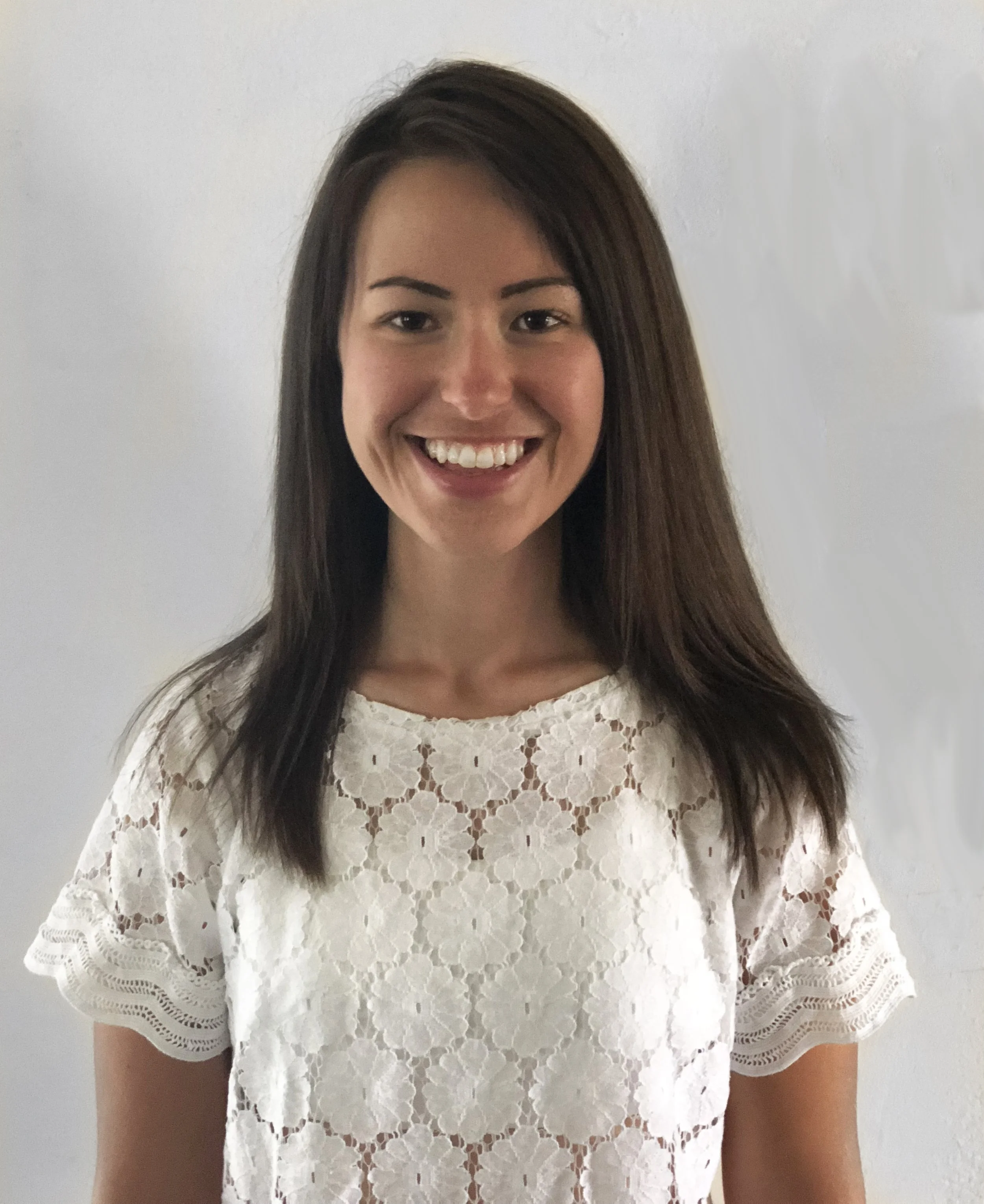 A young woman with long brown hair smiling, wearing a white lace top, standing against a plain white wall.