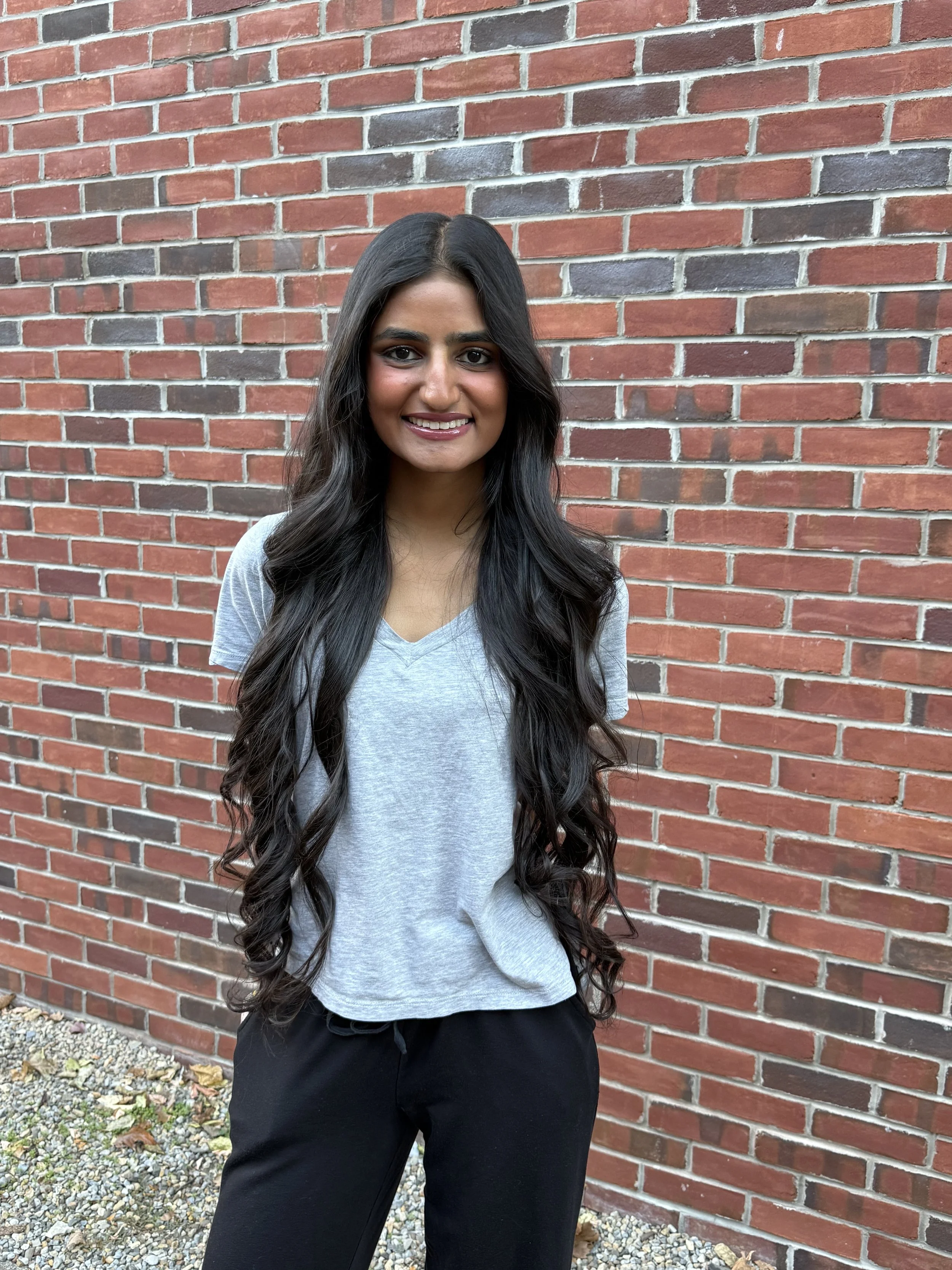 A woman with long, wavy black hair is smiling and standing in front of a red brick wall. She is wearing a light gray V-neck t-shirt and black pants.