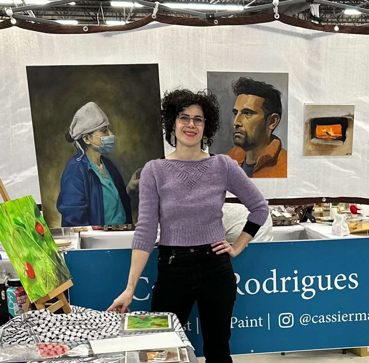 A woman with curly hair, glasses, and a purple sweater standing in front of art paintings at an art booth. The background includes portraits of a healthcare worker and a man, along with a small painting of a fireplace. Art supplies and paintings are on the table and easel.