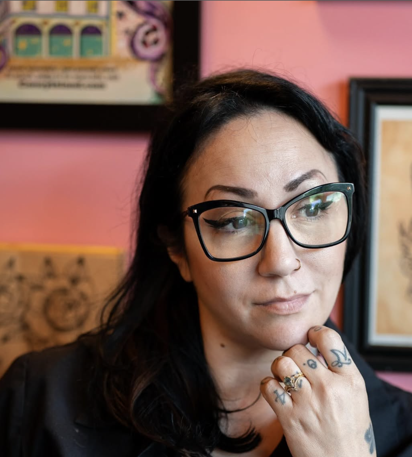 A woman with black hair, glasses, and tattoos touches her chin, posing thoughtfully in front of pink and framed artwork on a wall.