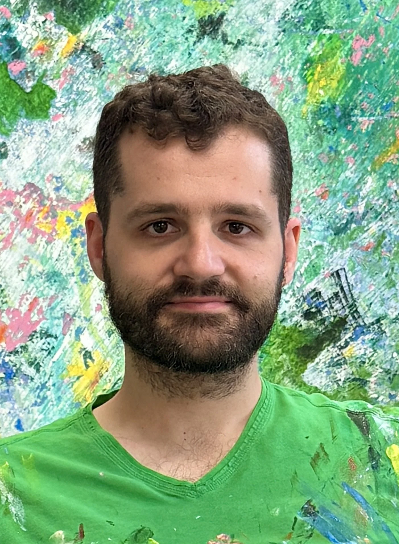 A man with a beard and curly brown hair, wearing a green shirt, standing in front of a multicolored abstract painting.