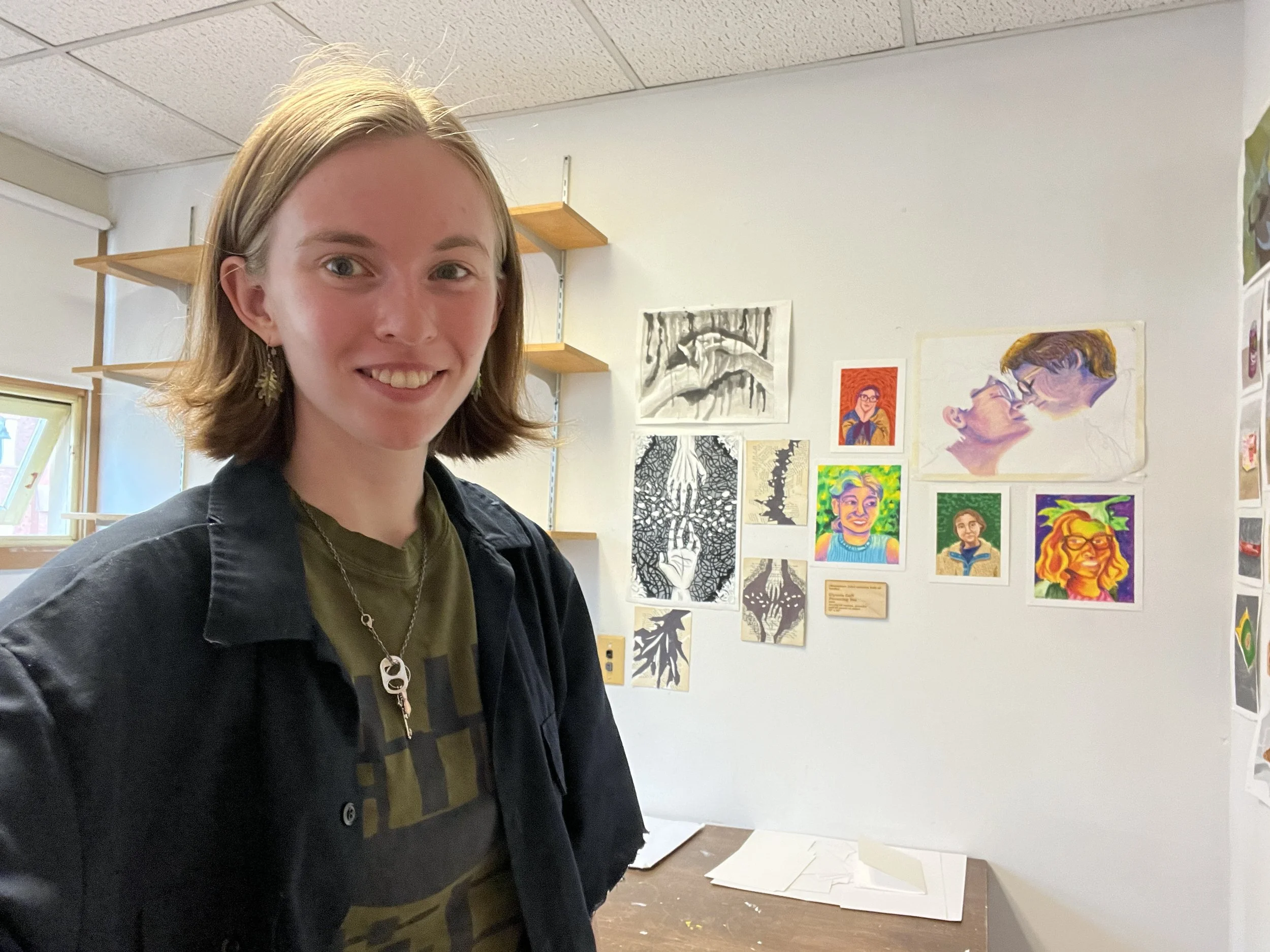 A young woman with shoulder-length light brown hair wearing a dark jacket and a green shirt, standing in an art studio with paintings and drawings on the wall behind her.