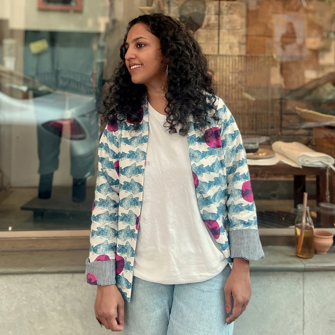 A woman with curly black hair and hoop earrings stands outside a store window, smiling and looking to her left. She is wearing a white T-shirt and a patterned blazer with pink, blue, and white colors. The background shows a display of towels and other items inside the shop.