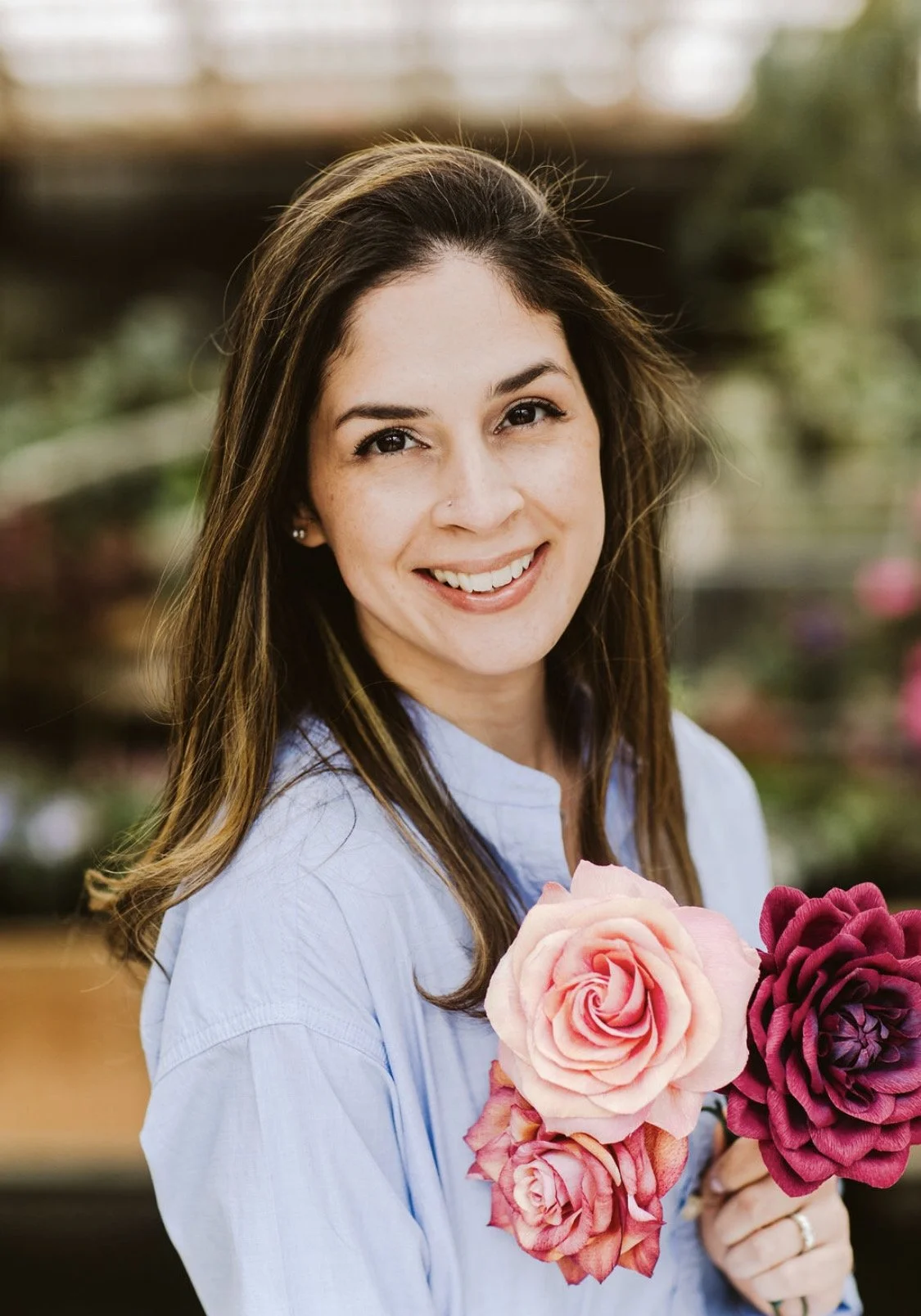 A woman with long brown hair smiling while holding pink and purple roses, wearing a light blue shirt, outdoors with blurred greenery in the background.