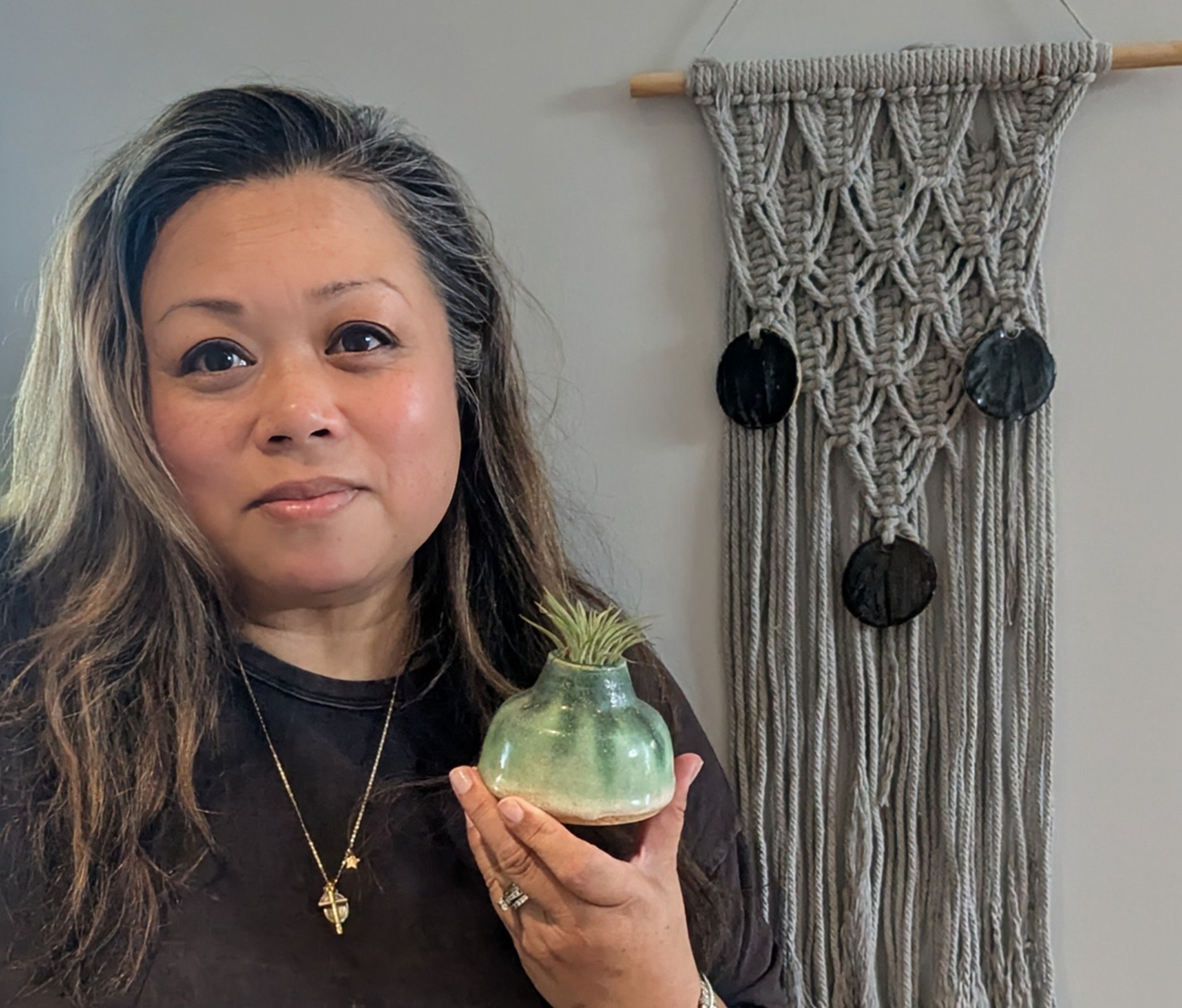 Woman with long dark hair holding a small ceramic vase with a green plant, standing in front of a macramé wall hanging.