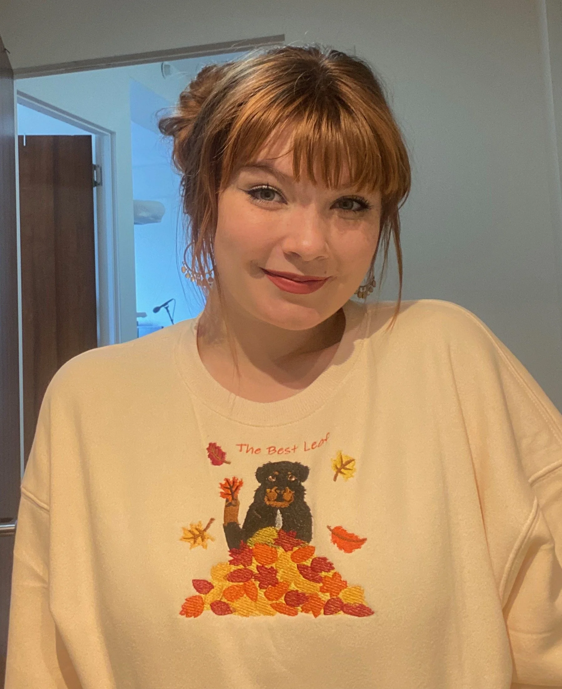 A woman with red hair and light skin wearing a cream-colored sweatshirt with an embroidered bear surrounded by fall leaves.