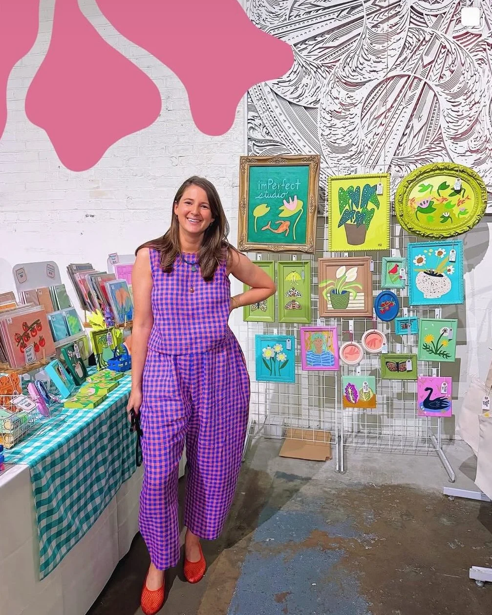 A woman standing in front of colorful artwork and greeting cards at an art and craft fair booth. She is wearing a purple checkered outfit and red shoes, smiling at the camera.