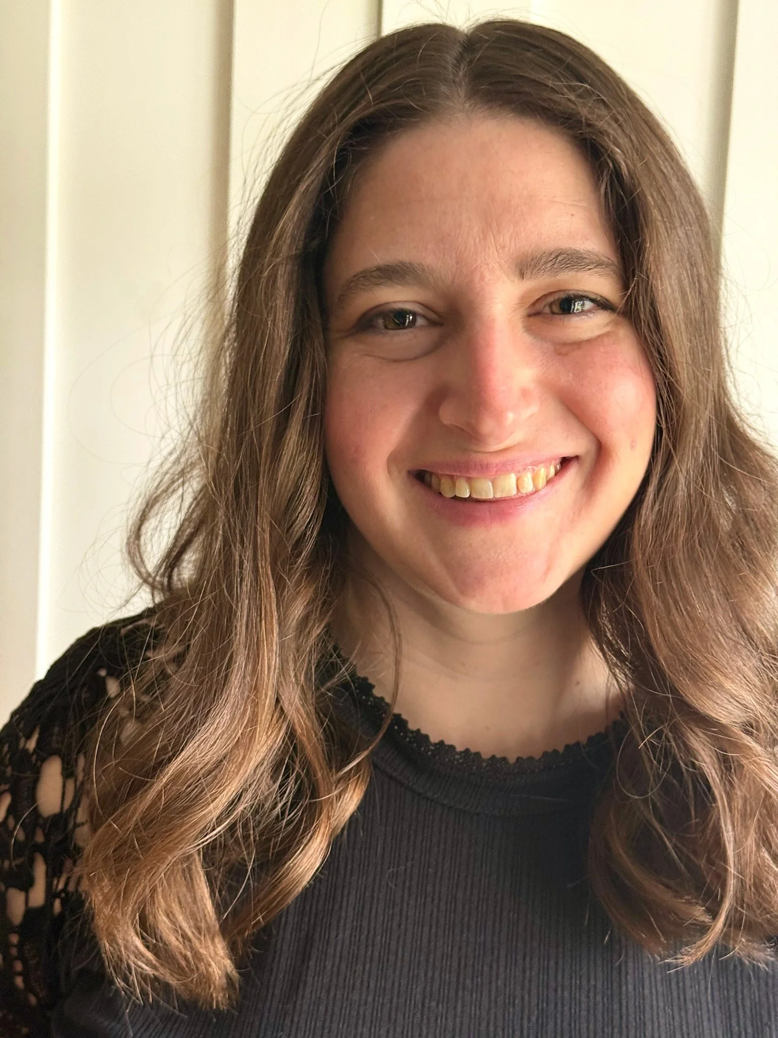 A woman with long, wavy brown hair smiling at the camera, wearing a black top with lace details.