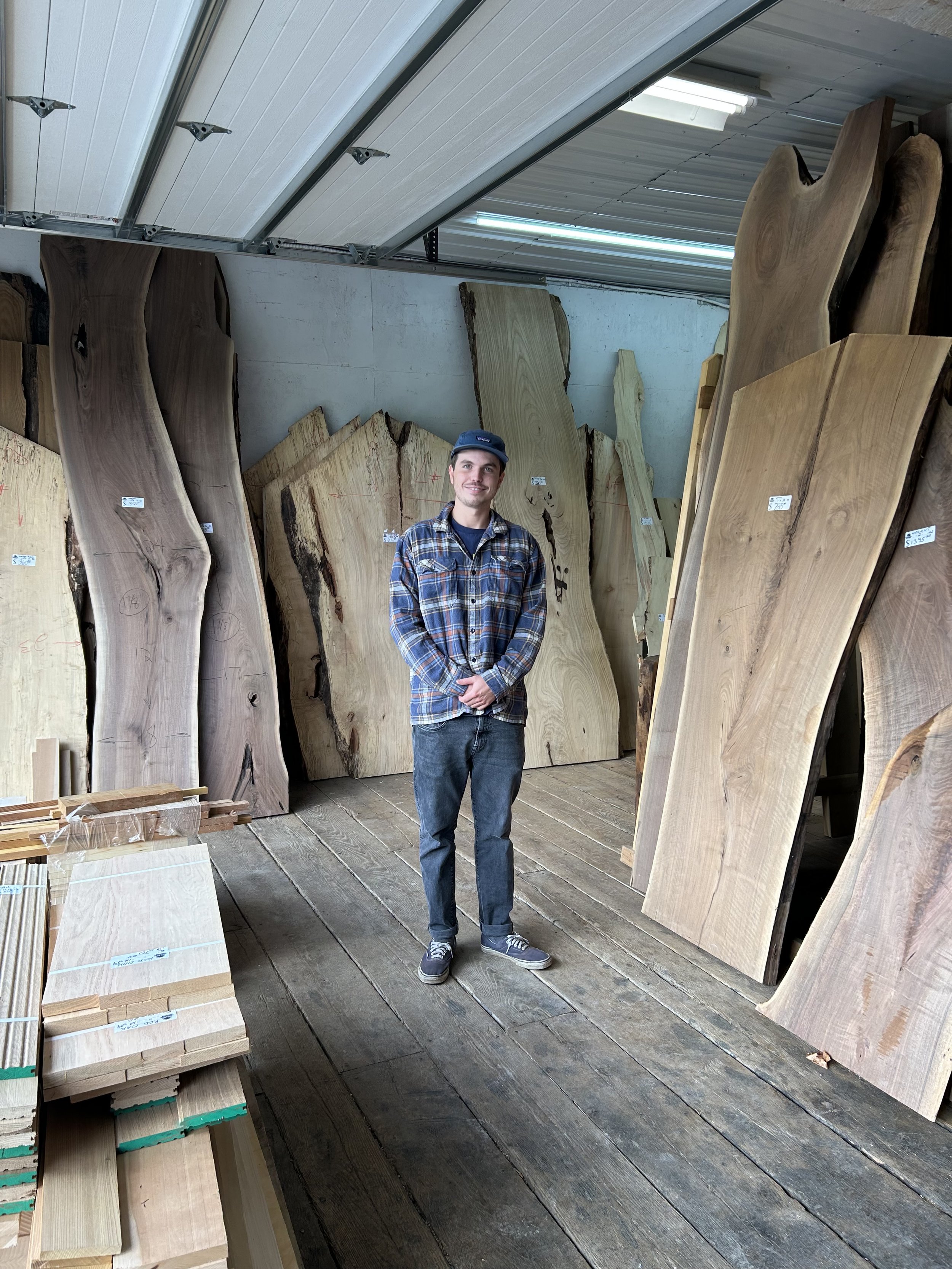 A young man in a plaid shirt and jeans standing inside a wood store surrounded by large slabs of wood and lumber.