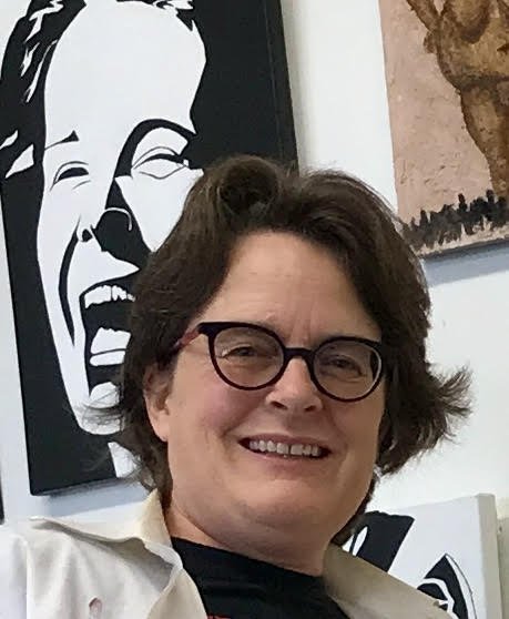 A woman with short brown hair and glasses smiling in front of artwork that features a smiling face and a brown-toned picture.