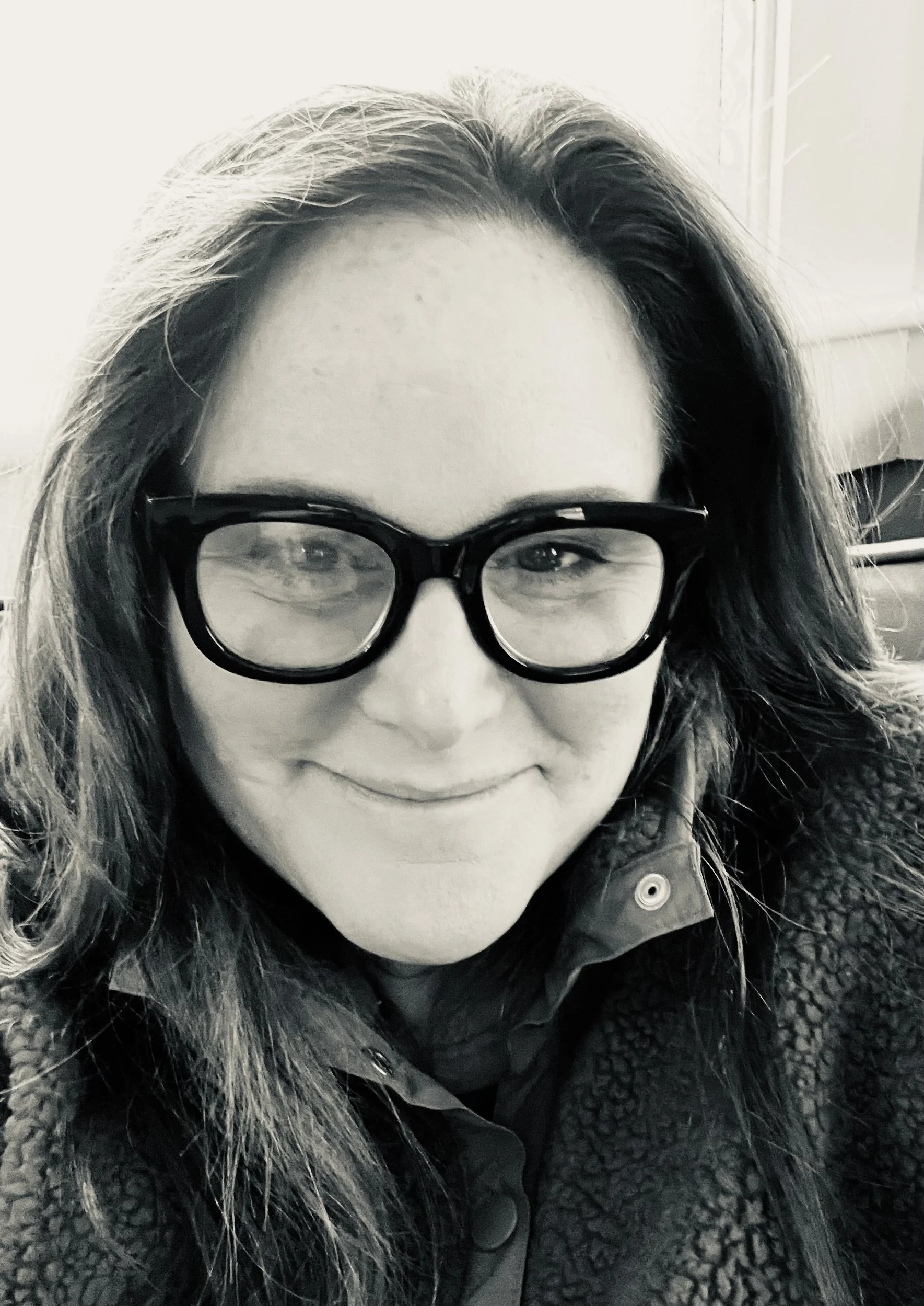 A woman with long hair, glasses, and a smile, wearing a textured jacket.