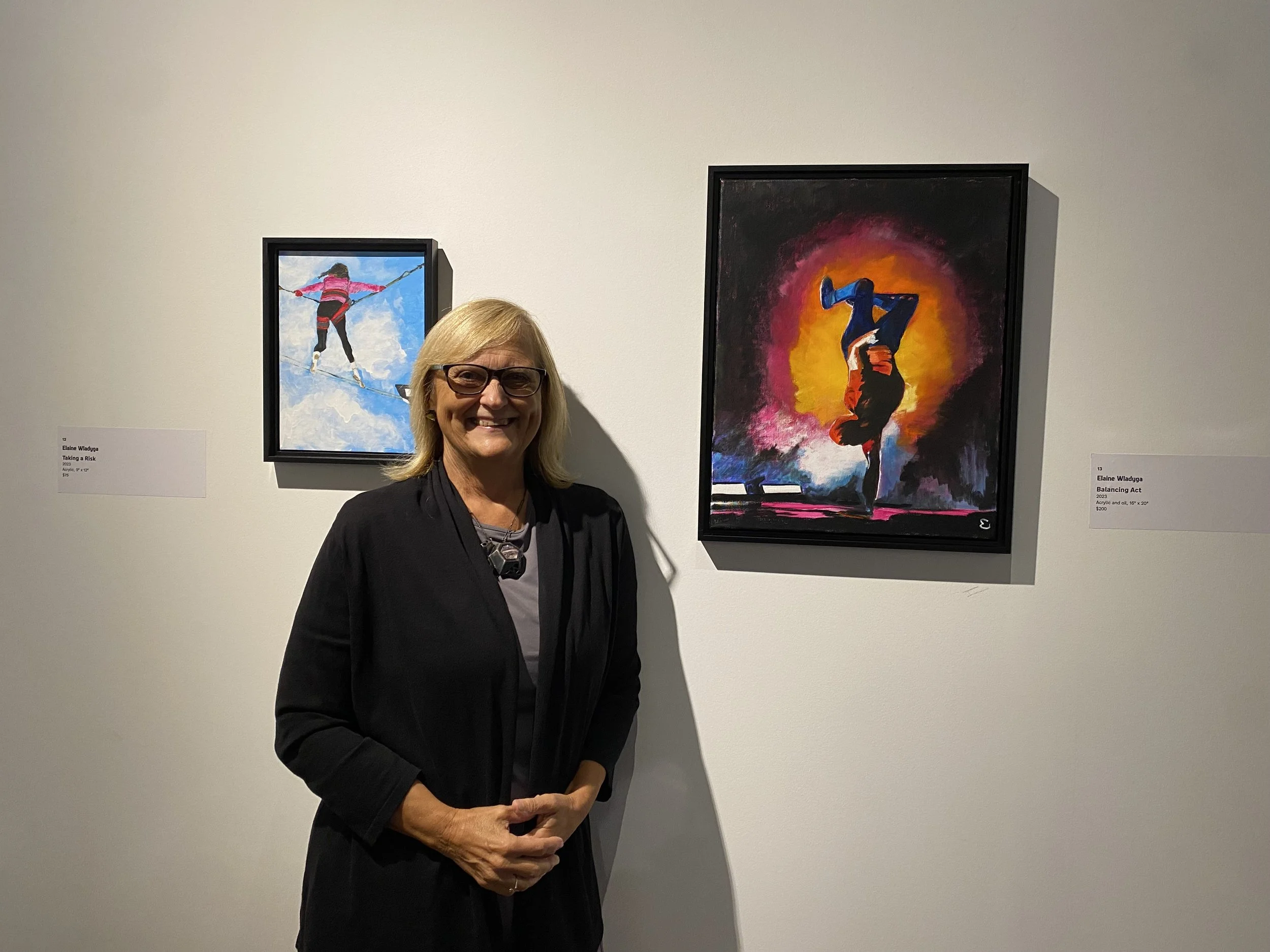 A woman with blonde hair and glasses standing in front of a white gallery wall, smiling. Two colorful paintings are displayed behind her, one of a person slacklining in the sky, and another of a person performing a handstand at sunset.