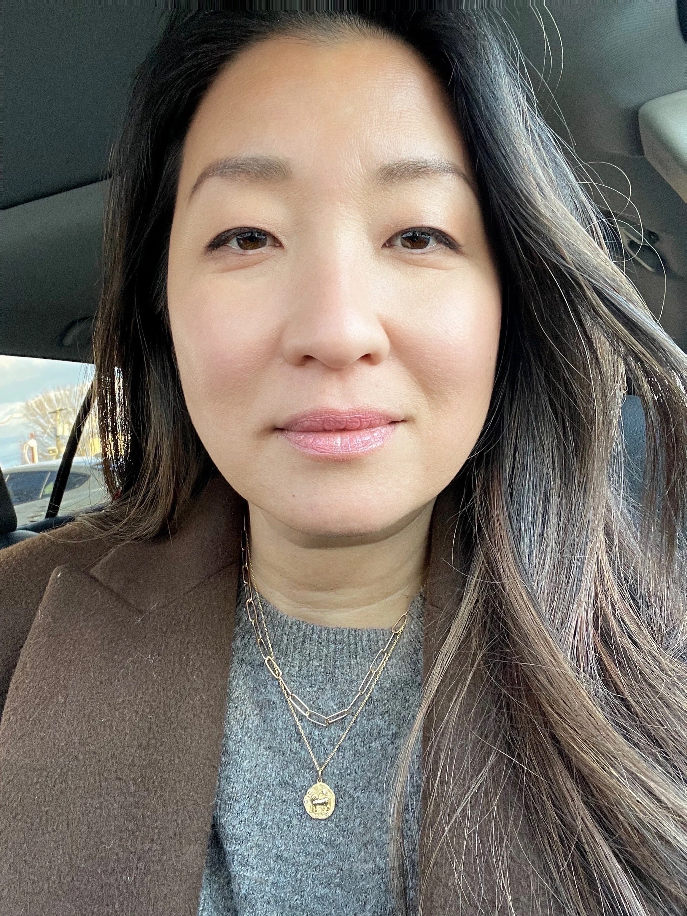 Close-up of a woman with long dark hair, wearing a gray sweater, gold necklaces, and a brown coat, sitting inside a vehicle.