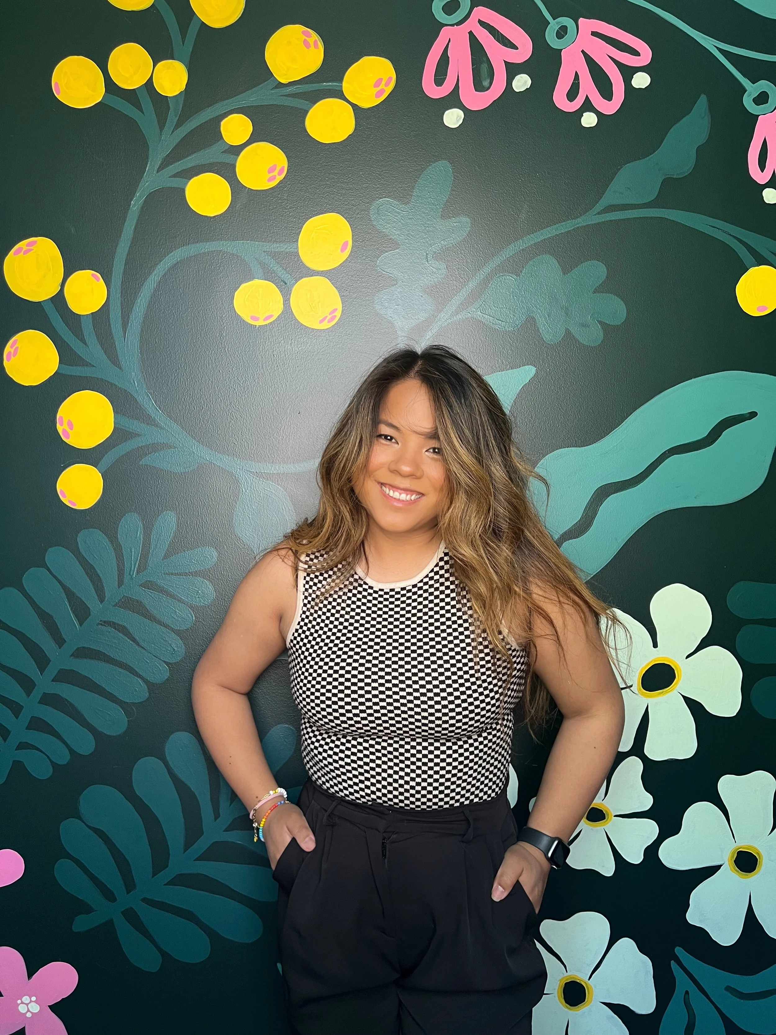 A woman with long wavy hair smiling and standing with hands in her pockets in front of a colorful mural featuring yellow berries, pink flowers, and green leaves.