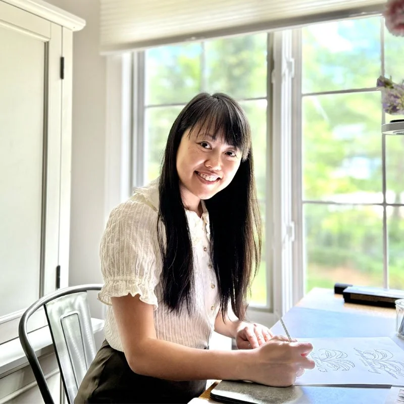 A woman with long dark hair, wearing a light-colored blouse, sitting at a desk by a large window, smiling and drawing in a sketchbook.