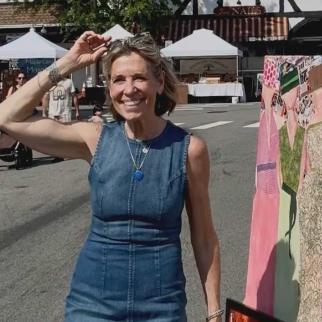 A smiling woman in a sleeveless denim dress, standing outdoors at a market with tents and people in the background.