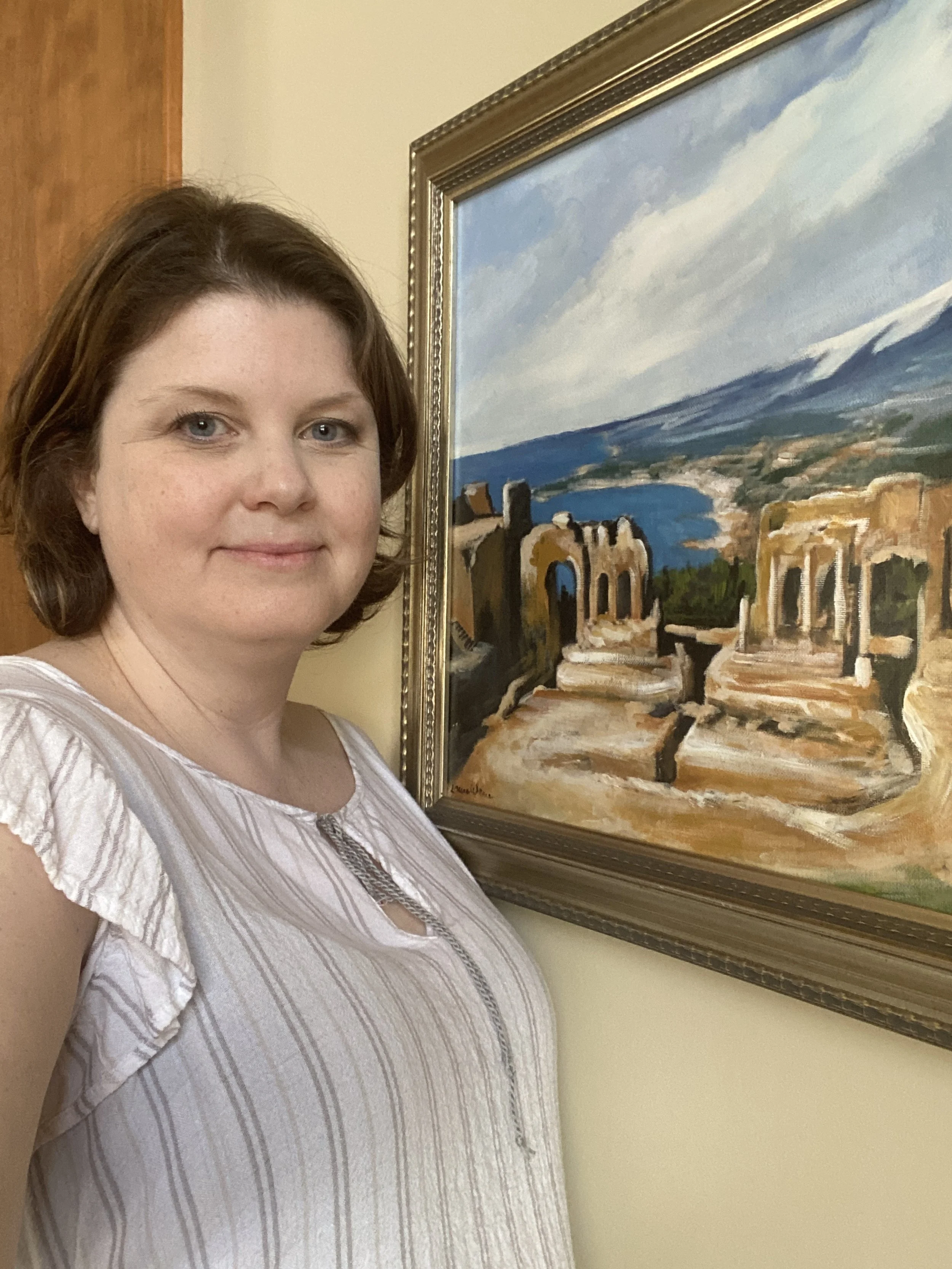 A woman with short brown hair standing next to a framed landscape painting of ruins and a lake, with mountains and sky in the background.