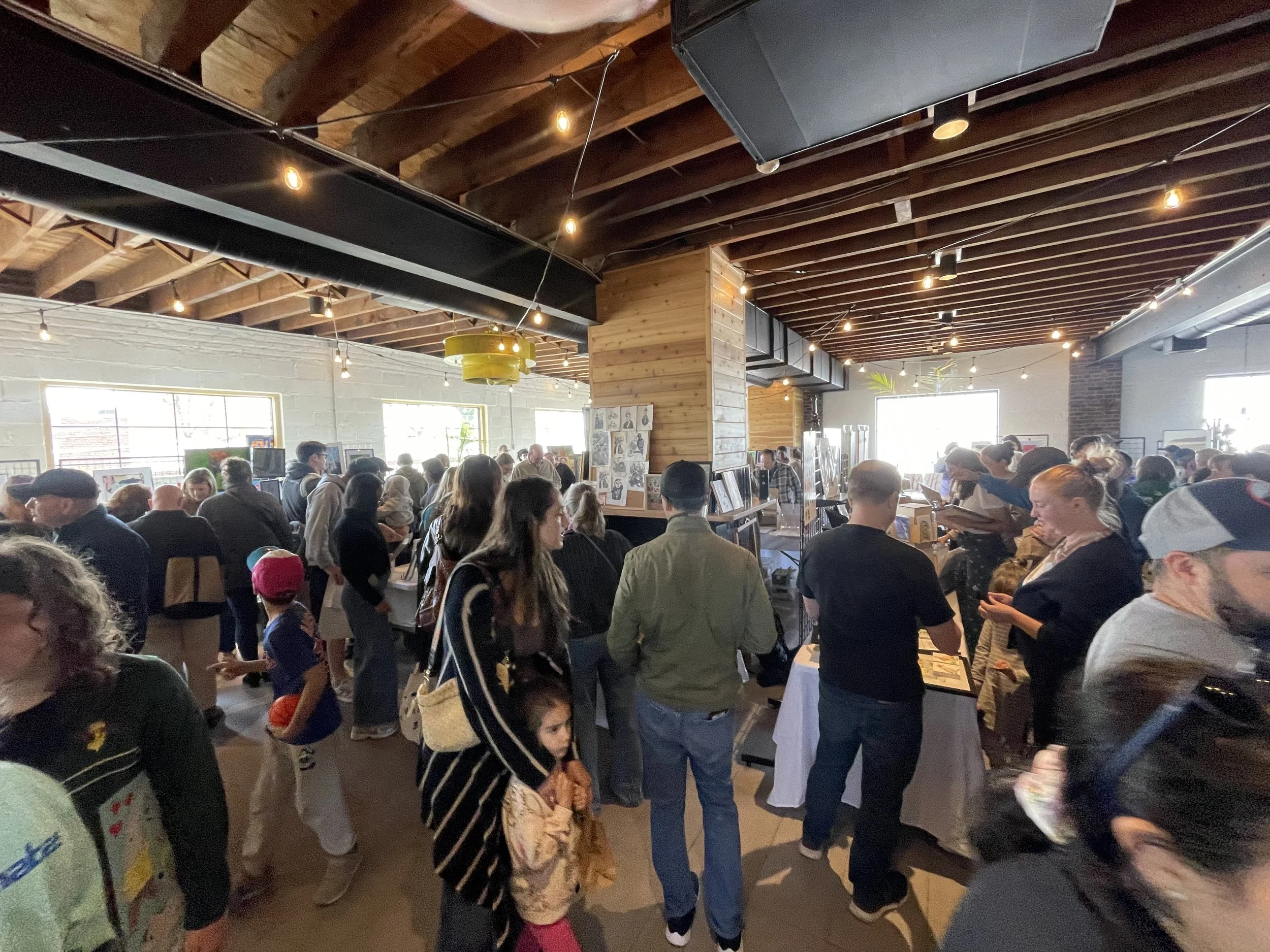 Crowd of people inside a large room with wooden ceiling beams, some using booths or tables, possibly at an art show or market.